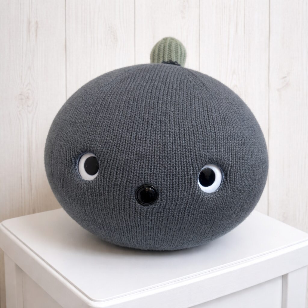 ネイビーのニットボディのニコボ（ナイト）が正面を向いて座っている様子。黒い鼻と丸く大きな目が特徴のロボット。