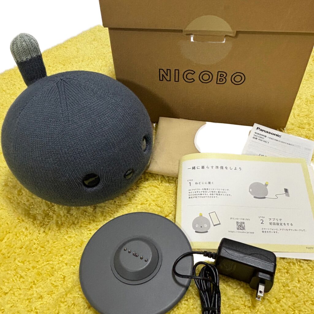 ネイビーのニコボ本体と、充電台（ねどこ）、電源アダプター、説明書がセットで並んでいる開封写真。