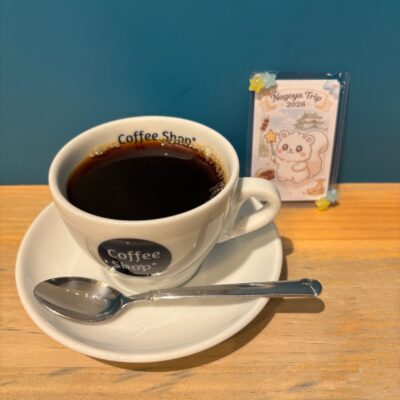 Coffee Shop の白いカップに注がれたホットコーヒー。奥に旅のルミリィの記念カードが飾られている。