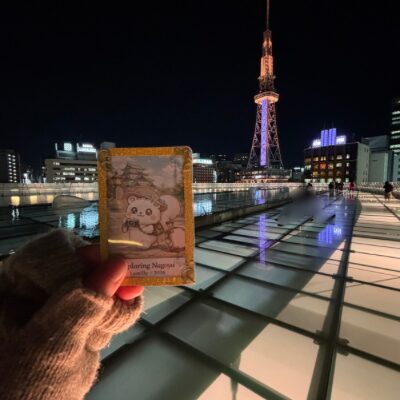 夜のオアシス21展望デッキで、名古屋テレビ塔を背景に、手袋をした手がブログのマスコットキャラクター・ルミリィのチェキを掲げている。ガラスの水盤に名古屋の灯りが美しく映り込んでいる。