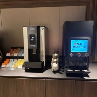 ホテルの宿泊者ラウンジにあるドリンクコーナー。ティーバッグが並んだ棚と、温冷どちらにも対応したドリンクサーバーが設置され、落ち着いた照明に包まれている。