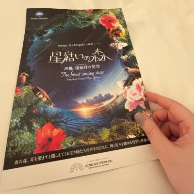ベッドの上で、コニカミノルタプラネタリウム『星結いの森』のチラシを手に持っている様子。柔らかな灯りと寝具が、旅の夜らしい穏やかな雰囲気をつくっている。