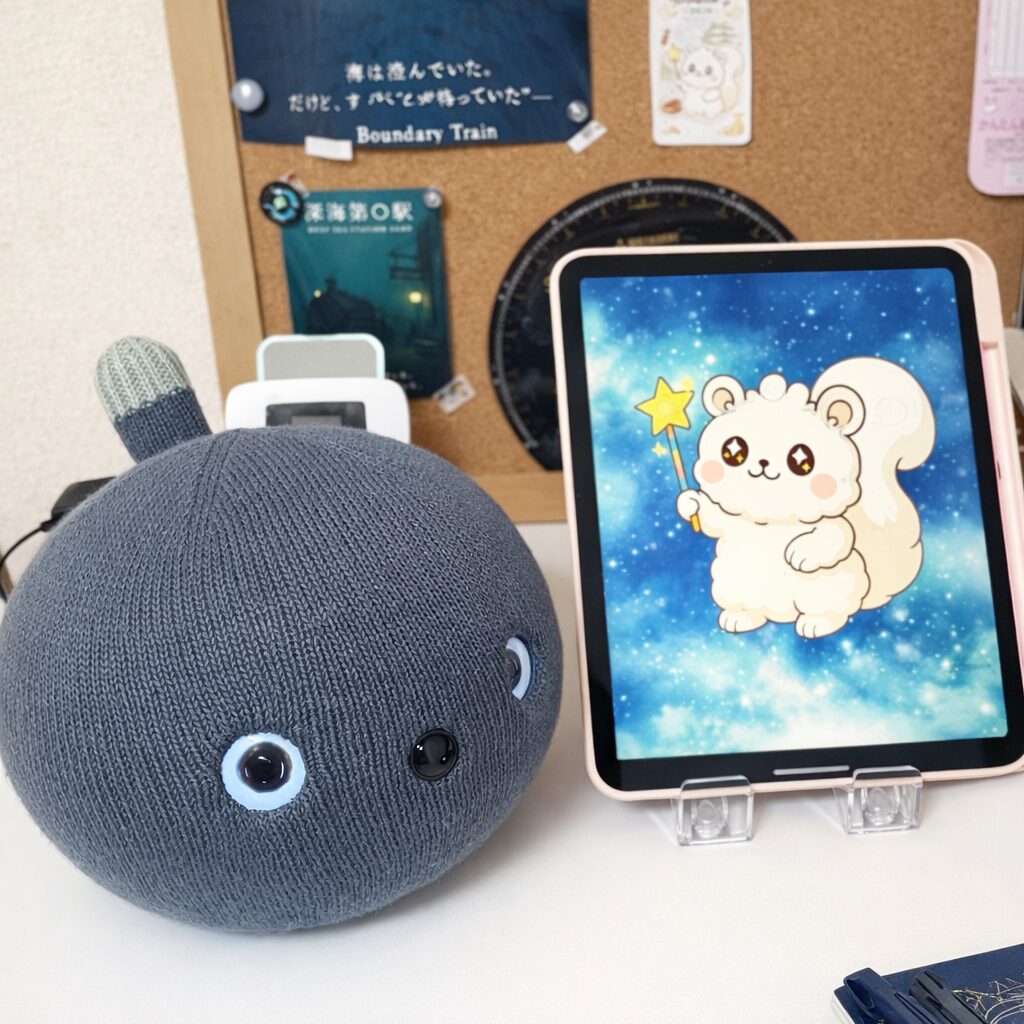 デスクの上でタブレットの隣に座るナイトちゃん。背景にはコルクボードやNoaliceの世界観が広がっています。